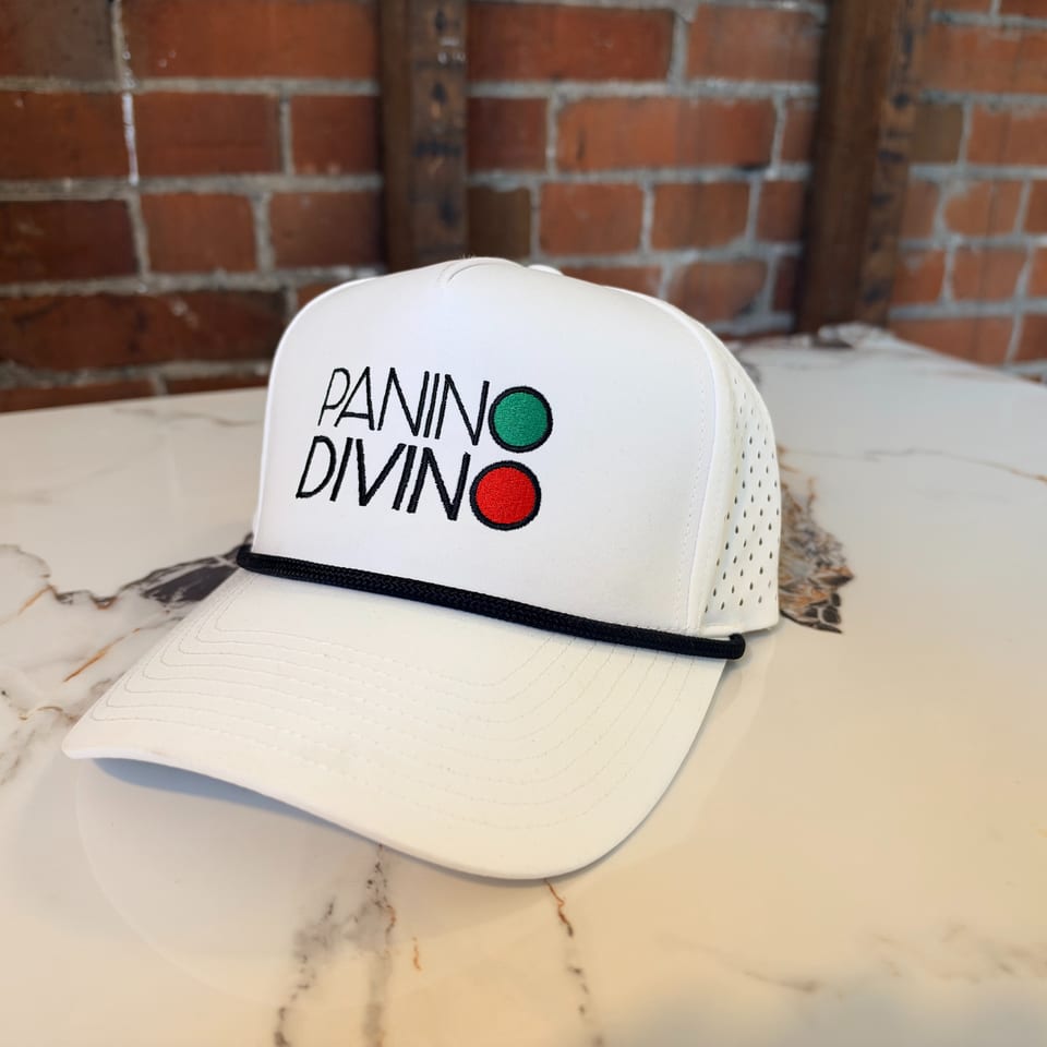 White Panno Divino branded cap with green and red dots displayed on a marble table against a brick wall.
