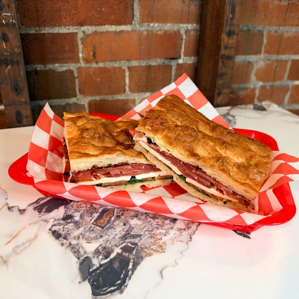 Two toasted deli sandwiches filled with sliced meats and cheese sit on red-and-white checkered paper in a red tray.